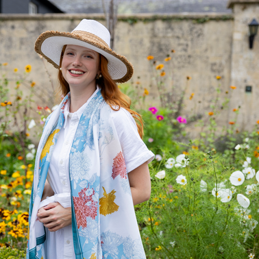 Woman wearing white straw hat and a blue and white silk-like lightweight scarf, in the garden. 