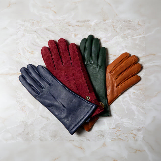 Four leather gloves in navy blue, burgundy, forest green, and tan, arranged in a fan shape on a light beige marble surface.