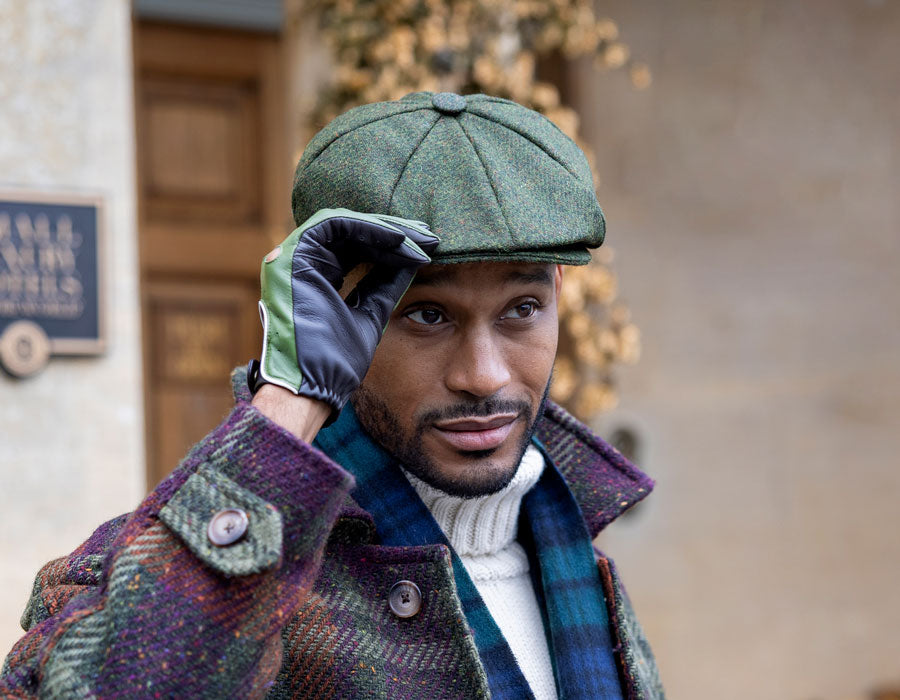 Man wearing a tweed flat cap in green with leather gloves in front of a building