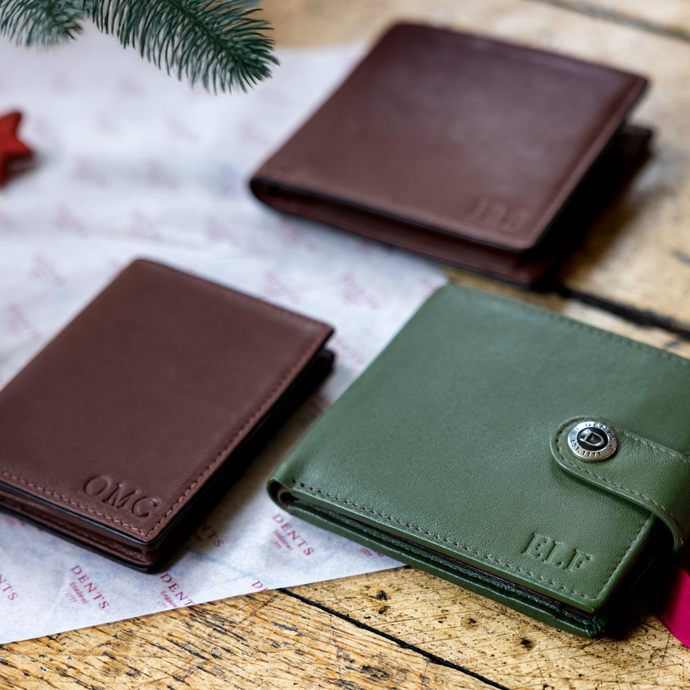 Two brown personalised men's leather wallets and one personalised green wallet on a wooden surface with a festive background.