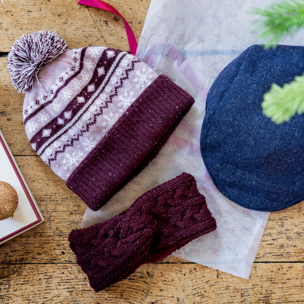 A blue men's tweed flat cap, women's purple fair isle knitted bobble hat and women's purple knitted headband against a festive background