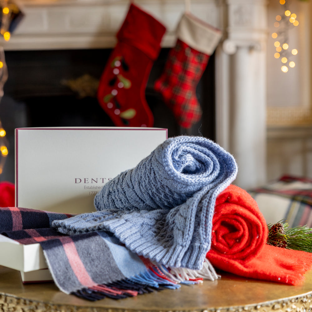 A blue cable knit scarf, red sequin scarf and checked cashmere scarf peeking from a Dents gift box on a table in front of a festive background