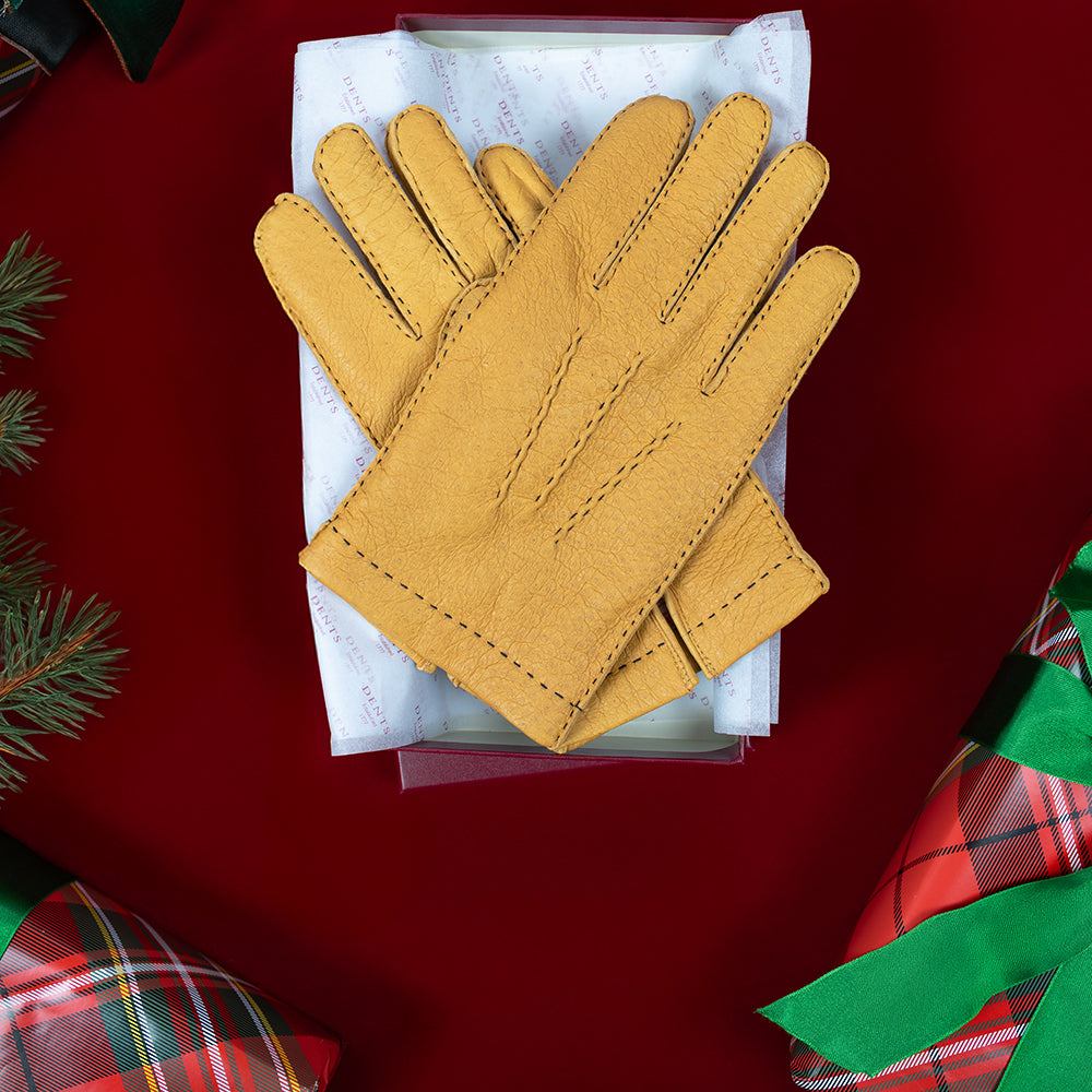 A pair of yellow peccary leather gloves with decorative contrast stitching, displayed on top of a Dents gift box against a dark red background with wrapped Christmas presents.