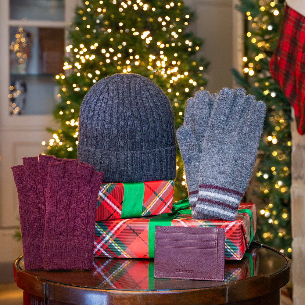 A holiday gift set featuring a men's charcoal grey ribbed knit beanie hat, grey knitted gloves, burgundy cable-knit fingerless gloves, and a burgundy leather card holder on top of wrapped Christmas presents on a wooden table with a blurred Christmas tree in the background.