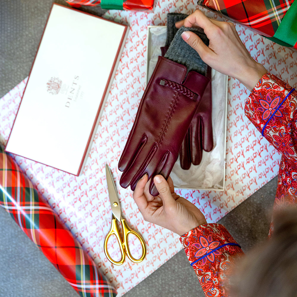 Close-up on woman's hands holding a pair of burgundy touchscreen leather gloves as she wraps them for Christmas in a Dents gift box, with gold scissors and red tartan wrapping paper.