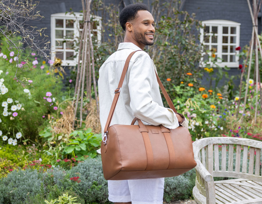 A man carries a caramel leather weekend bag over his shoulder in a garden setting with flowers and a bench.