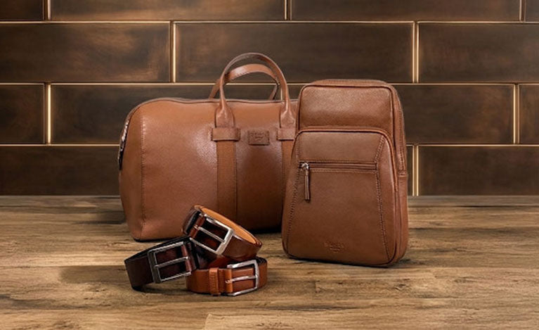 Brown leather briefcase, backpack, and belts on a wooden surface with a brick wall background.