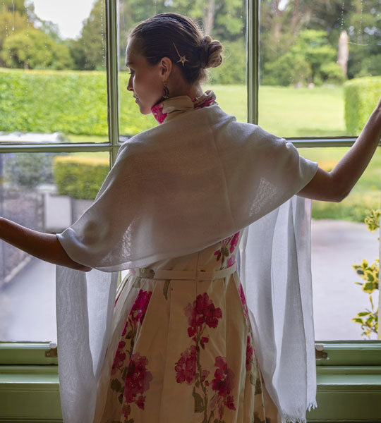 Woman wearing metallic silver lightweight scarf with floral evening gown standing by the window of a stately home