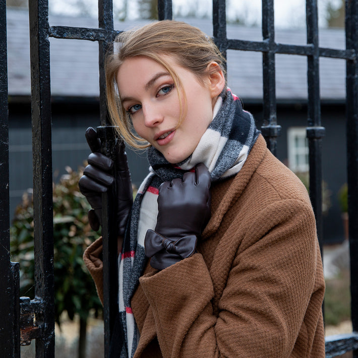 Woman wearing brown leather gloves with a bow with a checked scarf by a metal gate